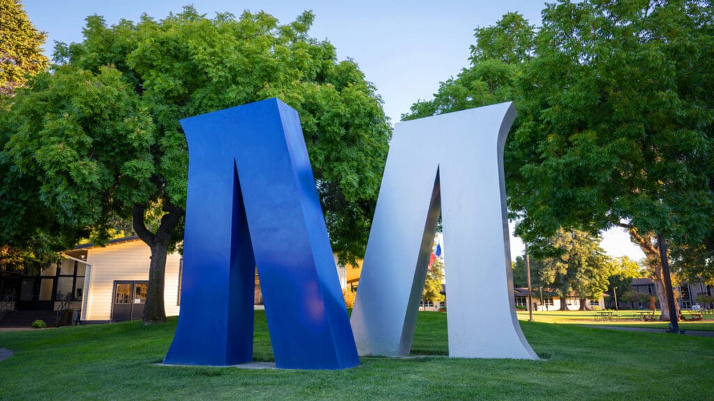 Menlo College’s large blue and white “M” sculptures stand on a grassy lawn surrounded by trees on the College’s Atherton campus.