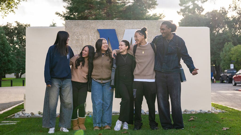 Six Menlo College students stand together smiling and laughing with their arms around one another in front of a large Menlo College campus monument sign, reflecting student camaraderie and community.