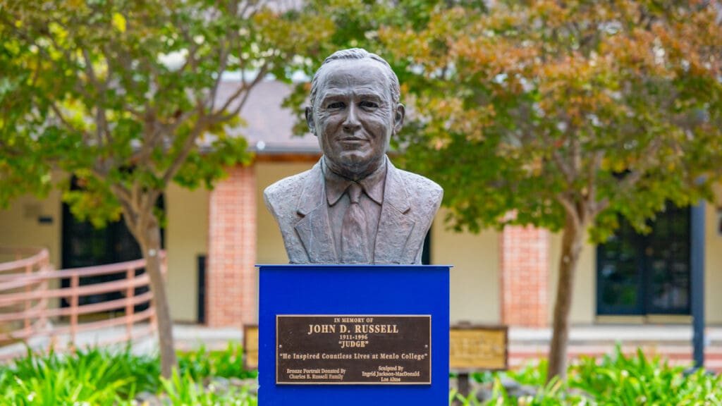 A bronze bust of John D. “Judge” Russell sits atop a blue pedestal with a commemorative plaque outside the Russell Center at Menlo College, surrounded by trees and landscaped greenery.
