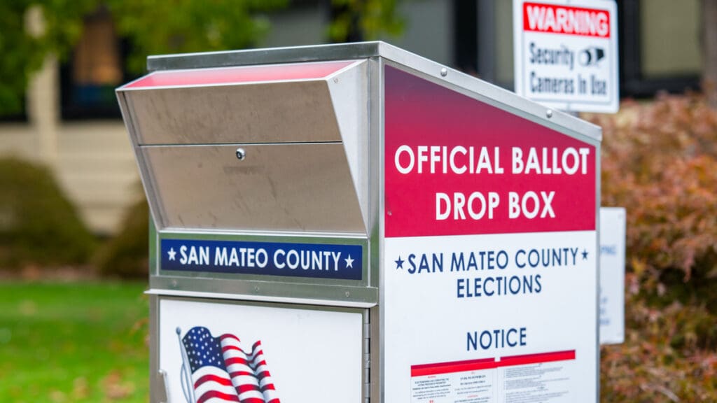 An official San Mateo County Elections ballot drop box stands near the entrance to Menlo College, featuring red, white, and blue signage, an American flag graphic, and instructions for returning completed ballots.