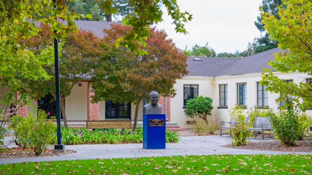 A bronze bust of John D. “Judge” Russell stands on a blue pedestal outside the Russell Center in El Camino Hall at Menlo College, surrounded by trees, landscaped gardens, and benches.